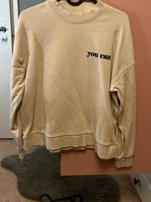 Beige sweatshirt med tryck 'you can' - En beige sweatshirt med rund hals och trycket 'you can' i svart på bröstet. Tröjan har långa ärmar och ribbade muddar vid hals, ärmslut och nederkant. sopass enkel och clean design som passar till  det mesta.