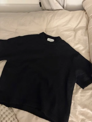 Svart stickad tröja från gina - En svart stickad tröja från gina med rund hals och korta ärmar. Tröjan har en loose passform och ribbade detaljer vid hals, ärmslut och nederkant. Perfekt för lager på lager under kyligare dagar.