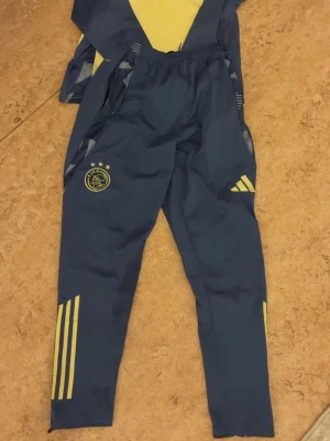 Blå/gul ajax tracksuit -  blå träningsbyxor från Ajax med gula detaljer och Adidas-logga, men inga skor finns med.