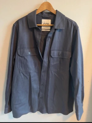 Mörkblå overshirt från COS - Snygg mörkblå overshirt från COS i regular fit. Jackan har två stora bröstfickor med knappar, klassisk krage och knäppning framtill. Materialet är bomull och ger en clean och stilren look. Perfekt att slänga över en t-shirt för en chill vibe.