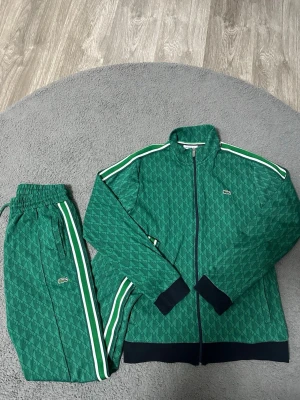Grönt set  från Lacoste - Säljer ett par gröna mjukisbyxor från Lacoste med vitt och grönt sidoband, elastisk midja och det klassiska krokodilmärket broderat på benet. Byxorna har ett diskret geometriskt mönster och är gjorda i mjukt material som är skönt att ha på sig. Med kommer tröjan! 