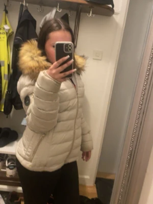 Beige pufferjacka med pälsluva - Säljer en beige pufferjacka med stor huva och fluffig fuskpäls i bruna toner. Jackan har dragkedja framtill och två snedställda fickor med dragkedja. Perfekt för kalla dagar och riktigt mysig att bära. Klassisk quiltad design och långärmad modell.