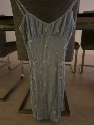 Blommig sommarklänning - Supersöt blå klänning med smala axelband och blommigt mönster i vitt och gult. Klänningen har en figurnära passform och är sydd i ett lätt, ribbat material. Perfekt för varma dagar och ger en fräsch vibe med sina små blommor och tunna ränder.