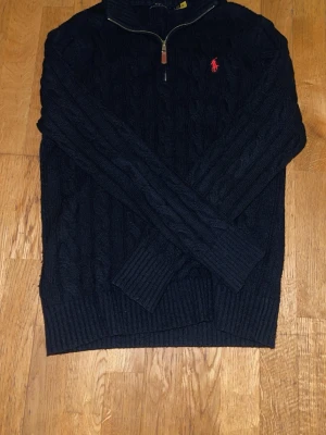Svart stickad tröja Polo Ralph Lauren - Kabelstickad mörkblå tröja från Polo Ralph Lauren med halv dragkedja och röd broderad logga på bröstet. Tröjan har lång ärm, ribbade muddar och hög krage. Perfekt för kyliga dagar och snygg till jeans eller chinos.