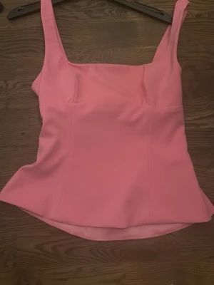 Rosa topp med rosett från Zara - Supersnygg rosa topp från Zara med breda axelband och en stor rosett i ryggen. Toppen har figurnära passform och är tillverkad i ett slätt, lätt material. Perfekt för dig som vill sticka ut med en feminin och trendig look.