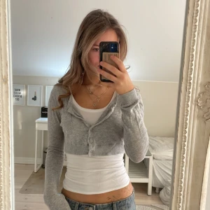 Grå croppad kofta med knappar - Säljer en ljusgrå croppad kofta med rund hals och långa ärmar. Koftan har ribbade muddar och knäppning framtill med fem knappar. Perfekt att slänga över ett linne eller t-shirt för en chill eller put together look.