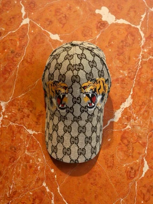Gcci keps med tigertryck - Säljer en snygg Gcci keps i beige med klassiskt GG-mönster och broderade tigrar framtill. Kepsen har gröna och röda ränder på sidan samt justerbart spänne bak med läderdetalj där det står Gcci. Perfekt accessoar för att sticka ut.