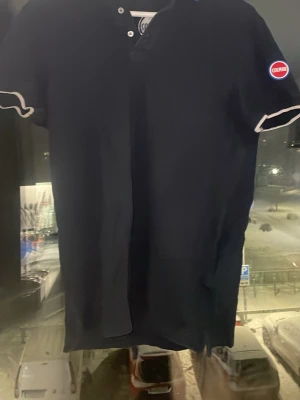 Mörkblå pikétröja från Colmar - Mörkblå pikétröja från Colmar Originals med korta ärmar och vit kant vid ärmslut. Tröjan har knappar vid halsen och ett broderat Colmar-märke på ena ärmen. Kragen har en snygg detalj med röd, vit och blå rand.