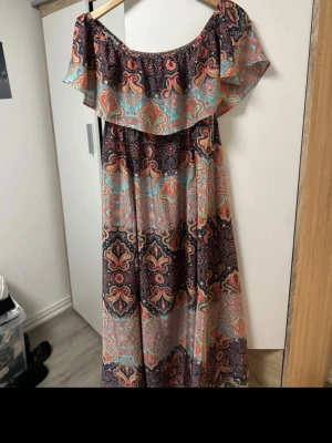 Mönstrad långklänning med volang - Färgglad långklänning med paisleyinspirerat mönster i blått, rött, orange och svart. Offshoulder-modell med bred volang upptill och lös passform. Perfekt för sommarens varma dagar. Materialet känns lätt och luftigt, troligen polyester eller bomull.