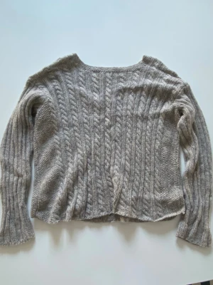 Grå kabelstickad tröja - Mysig grå kabelstickad tröja med lång ärm och rund hals. Tröjan har ett klassiskt flätmönster samt små blå detaljer. Egen-stickad och passar XS-M.