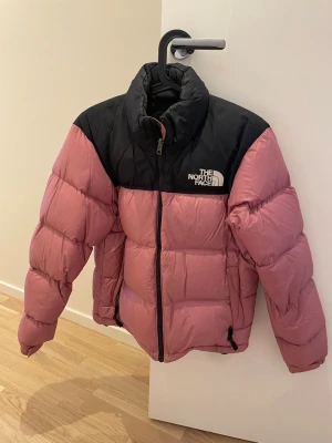 Rosa och svart pufferjacka North Face - Säljer en pufferjacka från The North Face i rosa med svart axelparti och hög krage. Jackan har dragkedja framtill och är vadderad för extra värme. Perfekt för kalla dagar och har en snygg, sportig vibe. Logga på bröstet och ryggen. Jackan är använd och har som enligt bild en del smink längst kragen, går att tvätta bort. 
