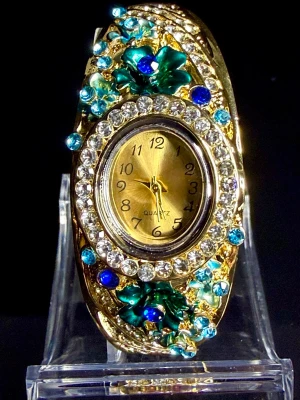 Pacific Blue Treasure  - Unik armbandsklocka i guld-look med blommiga detaljer och glittrande stenar i blått, grönt och turkost. Klockan har en oval urtavla och är dekorerad med gnistrande kristaller och metalliska blommor för en riktigt statement-look, funkar både till vardags och på fest!