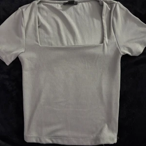 Beige topp med fyrkantig ringning - Snygg beige topp från New Yorker i storlek S. Toppen har en modern fyrkantig ringning och korta ärmar. Tillverkad i ett mjukt och stretchigt material som sitter skönt på kroppen. Perfekt att matcha med jeans eller kjol.
