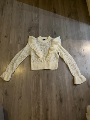 Vit stickad tröja med volangdetaljer - Superfin vit stickad tröja med volanger längs axlarna och ner över bröstet. Tröjan har ett snyggt hålmönster, långa ärmar med utsvängda muddar och ribbad kant nedtill. Perfekt för dig som vill ha något gulligt och trendigt i garderoben.