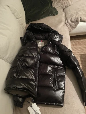 Svart pufferjacka från Moncler - Säljer en svart, glansig pufferjacka från Moncler med huva och dragkedja framtill. Jackan har två fickor med dragkedja och en Moncler-logga på ärmen. Perfekt för kalla dagar och riktigt snygg streetstil.