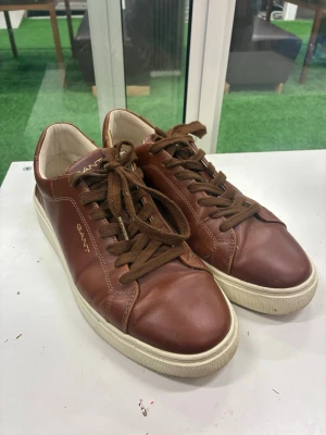 Bruna sneakers i skinn från Gant - Säljer ett par stilrena bruna sneakers från Gant i skinn med vita sulor och röda detaljer undertill. Skorna har klassisk snörning och rund tå. Perfekta för dig som vill ha en clean och tidlös look till vardags. Gant-logga i guld på sidan och plös.