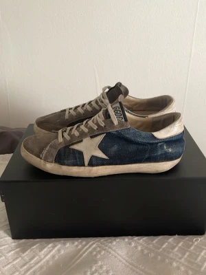 Golden Goose Superstar sneakers blå/grå - Ett par fina Golden Goose Superstar sneakers med blå denim på sidorna, grå mocka framtill och vita detaljer inkl den ikoniska stjärnan. Perfekta för dig som gillar en avslappnad och trendig look nu till våren!☀️Defekter: Sulan är lite trasig i hälen som de brukar bli på denna modell (Se sista bilden)✌🏼Storlek: 42 | Original skokartongen finns tyvärr ej kvar | Skriv gärna om ni har några frågor eller prisförslag!🎊