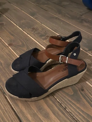 Marinblå kilklack sandaler med remmar  - Snygga marinblå sandaler med kilklack i rep och bruna justerbara remmar med spänne. Ovandelen är i tyg med korslagda band framtill och öppen tå. Perfekta för sommaren och ger en avslappnad men chic look. I mycket fint skick, använda fåtal gånger. Storlek 37