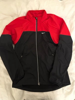 Nike Storm-FIT vindjacka röd/svart M - Nike Storm-FIT vindjacka i rött och svart med hel dragkedja framtill och två fickor med dragkedja. Jackan har hög krage, meshfoder och reflekterande detaljer. Finns även dragkedja vid armarna samt ficka baktill. Perfekt för blåsiga dagar och sportiga outfits.