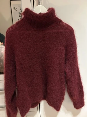 Mörkröd fluffig polotröja - Säljer en supermjuk och fluffig mörkröd polotröja med lång ärm i Kid mohair. Tröjan har en hög krage och är stickad i ett lurvigt material som känns riktigt cozy. Perfekt för kyliga dagar när du vill vara både varm och snygg. Storlek one size. 
