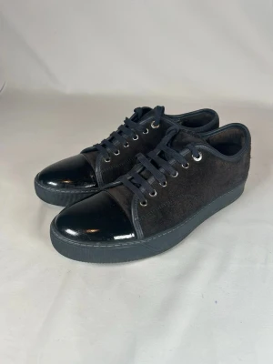 Svarta Lanvin DDB1 sneakers - (Nypris 4950kr)Snygga svarta sneakers från Lanvin med ovandel i mocka och glansig lackad tå. Skorna har snörning med svarta skosnören och metallöljetter. Sulan är svart och platt, vilket ger en clean och stilren look. Perfekta för dig som gillar en mix av material och en modern vibe.(Size 7uk men fit för 41/42)