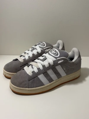Adidas Campus  - Säljer ett par grå Adidas Campus sneakers i mocka med klassiska vita ränder på sidorna och vit sula med gummidetalj. Skorna har vit snörning och rund tå. Perfekta för dig som gillar retrovibbar och streetstyle.