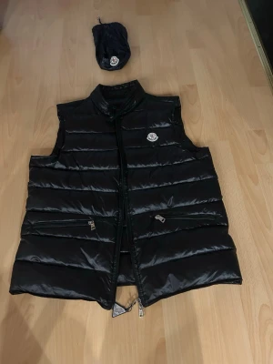 Svart dunväst från Moncler,  - Säljer en svart dunväst från Moncler med klassisk logga på bröstet. Västen har hög krage, två dragkedjeförsedda fickor och quiltad design. Tillhörande förvaringspåse medföljer. Perfekt för lager-på-lager och snygg till både hoodie och t-shirt.