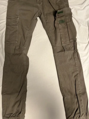 Beiga cargopants från Saligia - Säljer ett par beiga cargopants från Saligia med flera stora fickor på benen och klassisk cargostil. Byxorna har normal passform och är gjorda i bomull, perfekta för en chill och avslappnad look.