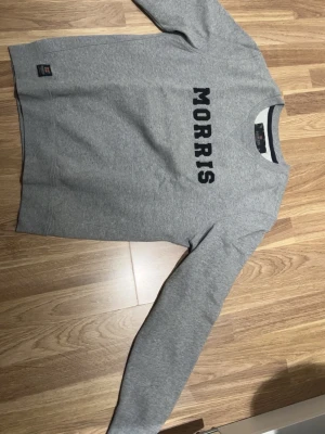 Grå sweatshirt från Morris, Small - Säljer en stilren grå sweatshirt från Morris i storlek Small. Tröjan har klassisk rund hals, långa ärmar och svart tryck med MORRIS på bröstet. Tillverkad i mjuk bomullsmix som är perfekt för både skola och chill. Enkel och clean design.
