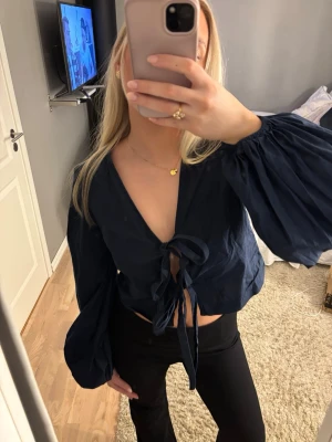 Mörkblå blus med knyt och ballongärmar - Säljer en mörkblå blus från Nelly med croppad passform, djup v-ringning och två knyt framtill. Blusen har långa ballongärmar med resår vid handlederna och är gjord i ett tunt bomullstyg. Perfekt för dig som gillar trendiga detaljer och en luftig känsla.