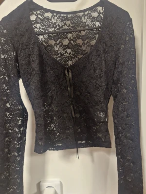 Svart spetsblus med snörning - Svart blus i genombruten spets med lång ärm och v-ringad hals. Framtill finns en snygg snörningsdetalj med satinband. Blusen är croppad och har ett blommigt spetsmönster som ger en cool och romantisk vibe. Perfekt för dig som gillar statement-plagg.