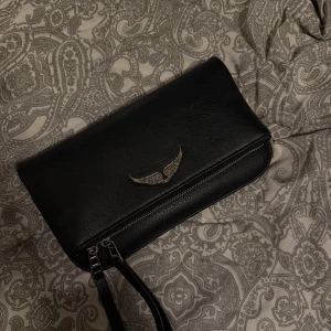 Äkta Zadig & Voltaire väska - Snygg svart axelväska i skinn från Zadig & Voltaire med silverfärgad kedja och coolt vingemblem framtill. Väskan har dragkedja både fram och bak samt avtagbar handledsrem. Perfekt storlek för det viktigaste och ger en edgy vibe till din outfit. Medföljer både långt och kort band. 