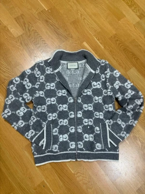 Grå stickad kofta från Gucci storlek M men sitter XS igentligen  - Säljer en grå och vit stickad kofta från Gucci med GG-mönster över hela plagget. Koftan har dragkedja framtill, ribbade muddar och krage. Tillverkad i Italien och har en snygg, lite loose passform. Perfekt för dig som gillar lyxiga detaljer och streetstyle. Pris är ej hugget i sten!