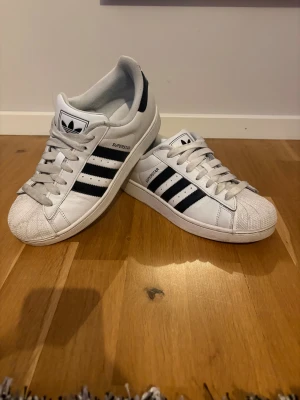 Adidas Superstar vita sneakers - Säljer ett par klassiska Adidas Superstar sneakers i vitt med svarta ränder och logga på hälen. Skorna har snörning, rund tå och den ikoniska snäckframsidan. Tillverkade i skinn med syntetdetaljer. Perfekta för dig som gillar retrostil och streetwear. Box ingår, skriv vid frågor!