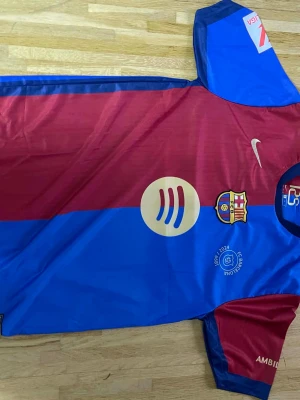 FC Barcelona Lamine Yamal matchtröja - Säljer en FC Barcelona matchtröja med Lamine Yamal #19 på ryggen. Tröjan är i klassiska blå och vinröda färger med Nike-logga, klubbmärke och sponsorer. Tillverkad i lätt polyester, perfekt för fotboll eller att bära som supporter.