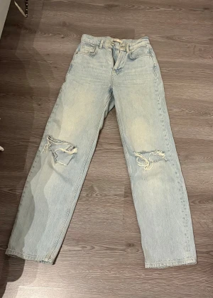 Ljusblå ripped jeans från Gina tricot - Ljusblå jeans från Gina tricot i rak modell med slitna detaljer och hål på båda knäna från petite-avdelningen. Klassisk femficksdesign, bälteshällor och dragkedja. Perfekt för en avslappnad och trendig look. 
