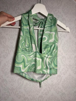 Grön swirl mönstrad linnetopp med knyt - Säljer en snygg grön linnetopp med swirl-mönster i olika gröna och vita nyanser. Toppen har djup v-ringning och knyt i nacken som ger en cool halterneck-look. Materialet är mjukt och glansigt, perfekt för att sticka ut. Passar dig som gillar färg och mönster! Mindre fläck på insidan 🫟 syns ej när du bär den. 