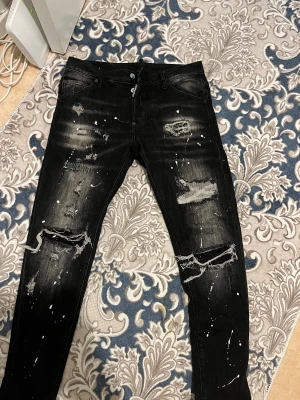 Svarta dsquared2 jeans helt nya! Storlek 30/32 - Svarta jeans med skinny fit, slitna detaljer och hål på knäna. Byxorna har vit färgstänkseffekt och en tvättad look som ger en riktigt edgy vibe. Klassisk femficksmodell och dragkedja. Perfekta för dig som gillar streetstyle.