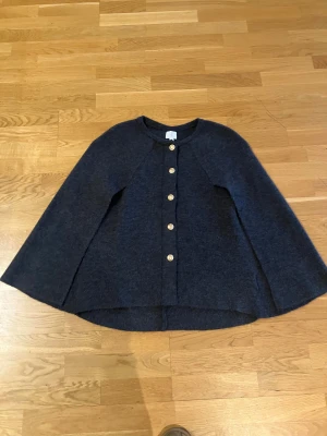 Mörkgrå kofta i cape modell med guldfärgade knappar - Snygg mörkgrå kofta med rund hals och sex guldfärgade knappar framtill. Koftan har en loose passform i cape modell och är tillverkad i ett mjukt, stickat material som känns skönt mot huden. Perfekt för dig som gillar stilrena och enkla plagg med en twist.