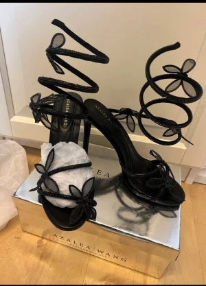 Svarta sandaler med höga klackar Azalea Wang - Svarta klackar från Azalea Wang med höga klackar och spiralformade remmar som snurrar upp längs benet. Dekorerade med fjäril detaljer i mesh och glittrande material. Öppen tå och platt sula framtill, perfekt för att sticka ut med en unik look.