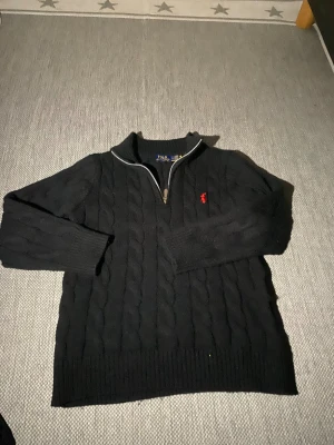 Svart stickad tröja Polo Ralph Lauren - Svart kabelstickad tröja från Polo Ralph Lauren med halv dragkedja och röd broderad logga på bröstet. Tröjan har lång ärm, ribbade muddar och hög krage. Perfekt för dig som gillar klassisk och stilren look.