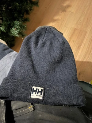 Mörkblå Helly Hansen mössa - Snygg mörkblå mössa från Helly Hansen med uppvikt kant och HH-logga framtill. Tillverkad av 99% återvunnen polyester, vilket gör den både mjuk och hållbar. Perfekt för kalla dagar och enkel att matcha med din stil.