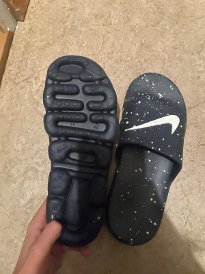 Svarta Nike sandaler med vita stänk - Säljer ett par svarta Nike sandaler med coolt vitt stänkmönster och stor vit swoosh-logga på ovansidan. Sandalerna har en platt sula med unik struktur undertill och är tillverkade i syntetmaterial. Perfekt för chill dagar eller efter träning.