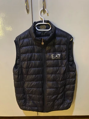 Svart dunväst från EA7 Emporio Armani - Snygg svart dunväst från EA7 Emporio Armani med quiltad design och hög krage. Västen har dragkedja framtill och logga på bröstet. Perfekt för lager-på-lager och ger en sportig vibe.
