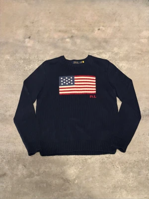 Mörkblå stickad tröja Polo Ralph Lauren - Snygg mörkblå stickad tröja från Polo Ralph Lauren med amerikansk flagga broderad på bröstet och RL-detalj i rött. Rund hals och ribbade muddar ger en klassisk look. Perfekt för dig som gillar preppy stil och vill sticka ut med en ikonisk design.