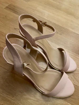 Ljusrosa sandaler med klack från T.PuSelle - Snygga ljusrosa sandaler från T.PuSelle med smal rem över tårna och justerbar rem runt vristen med guldfärgat spänne. Skorna har öppen tå, låg klack och en stilren design i syntetmaterial. Perfekta för att lyfta din outfit med en feminin touch.