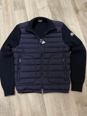 Mörkblå dunjacka från Moncler - Snygg mörkblå dunjacka från Moncler med quiltad front och stickade ärmar. Jackan har dragkedja framtill, två fickor med dragkedja och klassisk Moncler-logga på ärmen. Perfekt för kyliga dagar och ger en clean, sportig look.