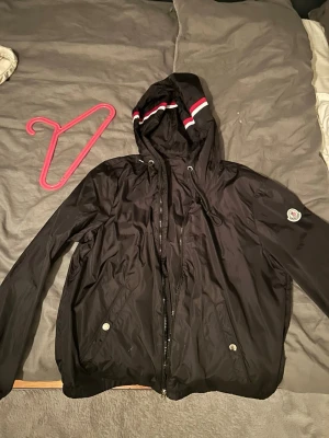 Svart Moncler vindjacka med huva - Svart vindjacka från Moncler med huva och snörning, dragkedja framtill och två fickor med tryckknappar. Huva har coola ränder i vitt och rött. Moncler-logga på vänster ärm. Jackan är tillverkad i lätt, glansig polyester som ger en clean och sportig vibe.