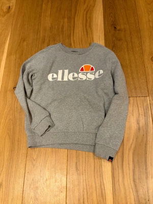 Grå sweatshirt från Ellesse - Snygg grå sweatshirt från Ellesse med stort vitt och orange logotryck på bröstet. Tröjan har rund hals, långa ärmar och ribbade muddar vid ärmslut och nederkant. Perfekt för dig som gillar sportig och avslappnad stil.