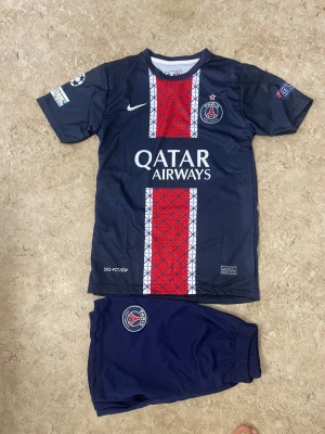 PSG fotbollströja och shorts Nike - Säljer ett Paris Saint-Germain matchställ med tröja och shorts från Nike. Tröjan är mörkblå med röda och vita detaljer, PSG-logga och Qatar Airways-tryck på bröstet samt D. Doué och nummer 14 på ryggen. Shorts i matchande mörkblå med PSG-emblem. Perfekt för fotbollsfans!